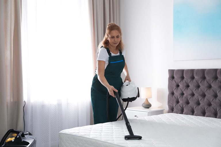 A woman is vacuuming the bed in her bedroom.